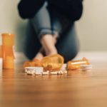 Orange prescription bottles spilled on a wooden floor with pills scattered; a person sits blurred in the background.