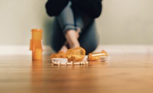 Orange prescription bottles spilled on a wooden floor with pills scattered; a person sits blurred in the background.