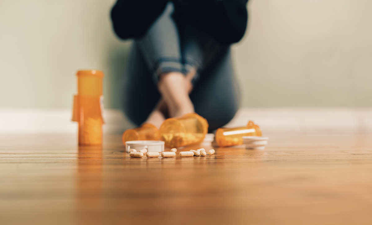 Orange prescription bottles spilled on a wooden floor with pills scattered; a person sits blurred in the background.