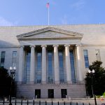 Neoclassical government building with tall marble columns and a triangular pediment, flag flying atop, steps leading to the entrance.