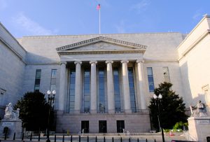 Neoclassical government building with tall marble columns and a triangular pediment, flag flying atop, steps leading to the entrance.