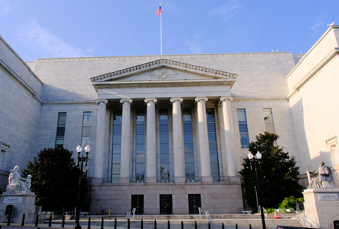 Neoclassical government building with tall marble columns and a triangular pediment, flag flying atop, steps leading to the entrance.